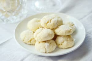 A white plate with a stack of round, cracked, pale Key Lime Cookies sits on a white tablecloth. The cookies appear soft and powdered, with a glass item blurred in the background.