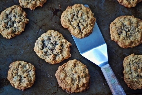 A metal spatula lifts a freshly baked Cowboy Cookie, loaded with oatmeal and chocolate chips, from a metal baking sheet surrounded by several more cookies.