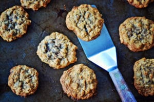 A metal spatula lifting a Cowboy Cookie from a baking sheet with several other cookies scattered around.