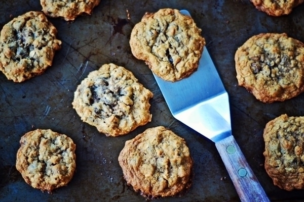 A metal spatula lifting a Cowboy Cookie from a baking sheet with several other cookies scattered around.