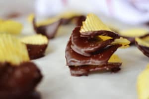 A close-up of Chocolate Covered Potato Chips shows ridged chips partially dipped in glossy chocolate, resting on parchment paper. Some chips are stacked, highlighting the sweet and salty combination.