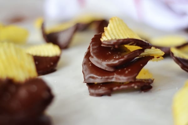 A close-up of Chocolate Covered Potato Chips shows ridged chips partially dipped in glossy chocolate, resting on parchment paper. Some chips are stacked, highlighting the sweet and salty combination.