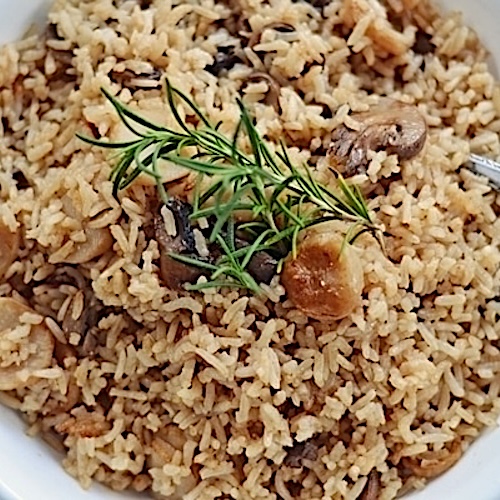 A close-up of a bowl of rice pilaf with sliced mushrooms, garnished with a sprig of fresh rosemary—this flavorful side dish pairs perfectly with Beef Rice Consomme.