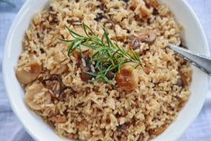A bowl of cooked rice mixed with sliced mushrooms and garnished with a sprig of fresh rosemary, served as a delicious side dish with a spoon resting on the side.