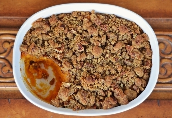A white oval dish filled with Sweet Potato Casserole, topped with a golden-brown, crumbly pecan streusel, with a serving missing from one side, revealing the rich orange sweet potato filling underneath.