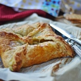 A golden-brown, flaky Baked Brie pastry square sits on parchment paper next to a silver knife and fork, with cookies and a red cloth visible in the background.