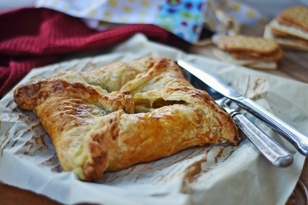 A golden-brown, flaky Baked Brie pastry square sits on parchment paper next to a silver knife and fork, with cookies and a red cloth visible in the background.