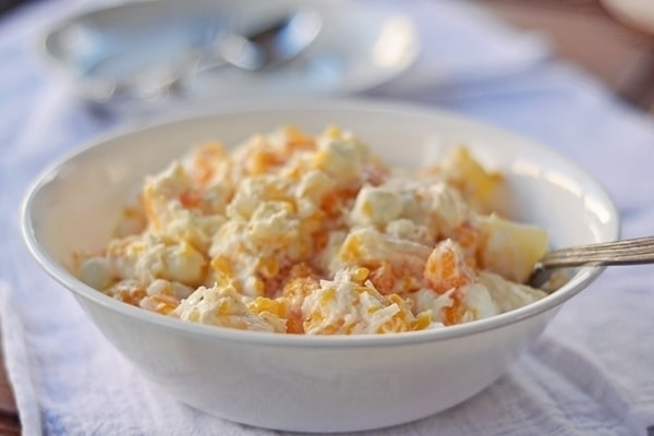 A white bowl filled with Russian salad made of chopped potatoes, carrots, peas, and mayonnaise, reminiscent of an Ambrosia 5 Cup Salad, with a spoon resting inside. The bowl sits on a white cloth with blurred plates and spoons in the background.