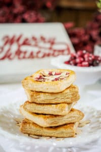 A stack of flaky, square Cranberry Cream Cheese Pastries topped with almonds and fruit sits on a white plate. In the background, there’s a bowl of cranberries and a festive Merry Christmas sign.