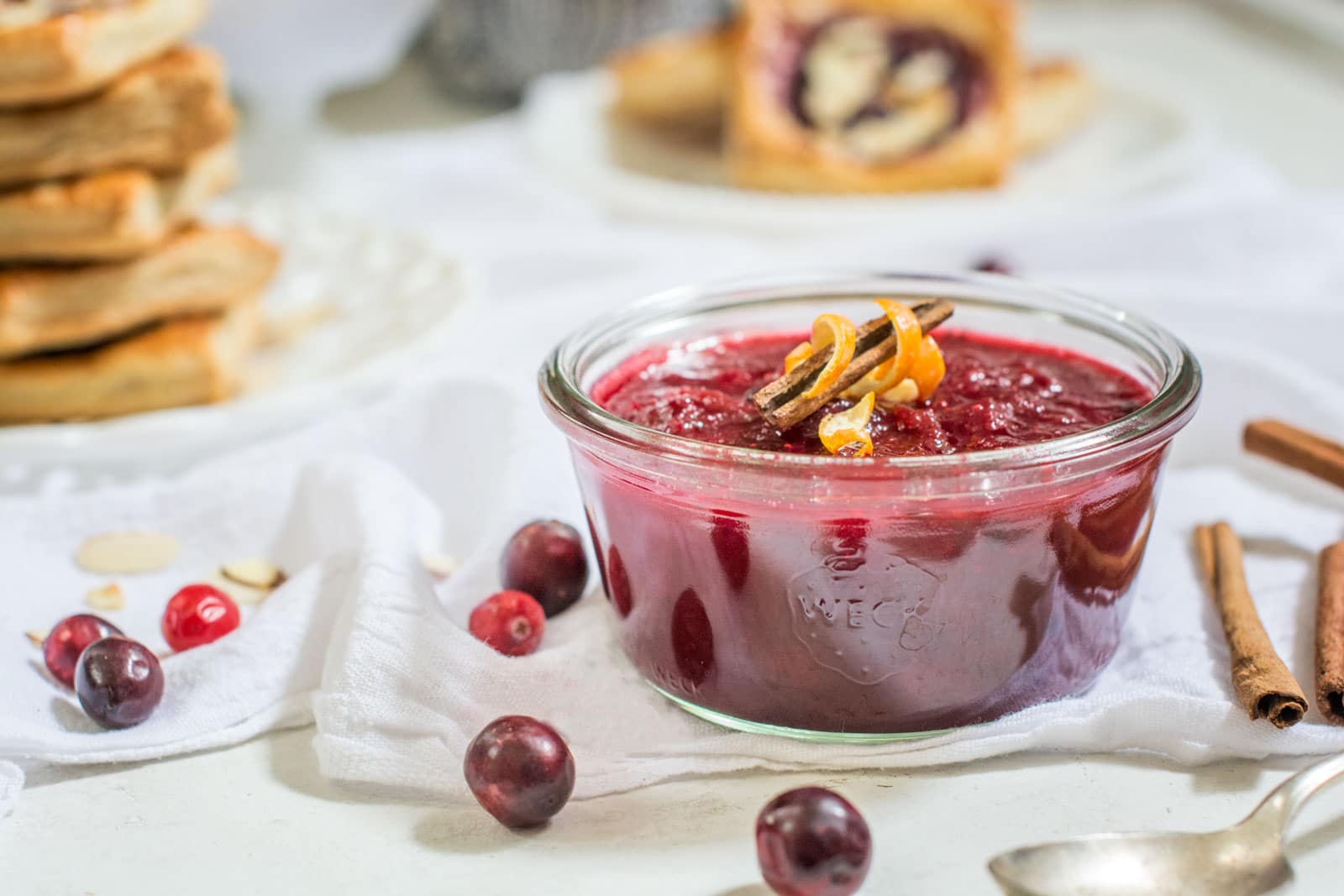 A glass bowl of Cranberry Sauce garnished with orange peel and cinnamon sticks is surrounded by fresh cranberries and pastries on a white tablecloth.