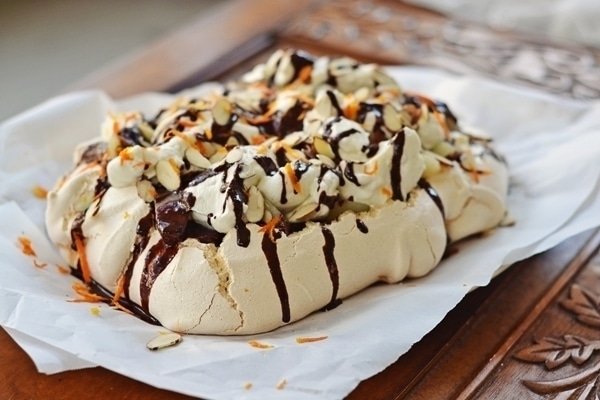 A homemade Pear and Chocolate Pavlova topped with whipped cream, sliced almonds, and drizzled with chocolate sauce sits on parchment paper atop a wooden surface.