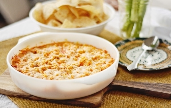 A baked cheesy Buffalo Chicken Dip in a white casserole dish is placed on a wooden board, with a bowl of tortilla chips and a plate in the background, ready for serving.