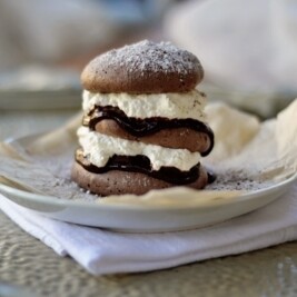 A Chocolate Pavlova dessert with layers of chocolate cookies, whipped cream, and chocolate filling is stacked on a white plate lined with parchment paper, dusted with powdered sugar. The plate rests on a folded napkin.