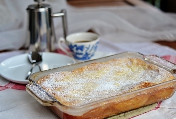 A glass baking dish filled with a golden Cream Cheese Coffee Cake, dusted with powdered sugar, sits on a table alongside a cup of coffee, a saucer, and a metal coffee pot in the background.