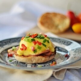 A plate with classic Eggs Benedict on an English muffin, topped with creamy hollandaise sauce, diced red and green peppers, and sliced ham, set on a patterned plate with vegetables blurred in the background.
