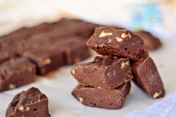 Close-up of several pieces of foolproof fudge with chopped nuts, stacked and arranged on a sheet of parchment paper, with more fudge pieces in the blurred background.
