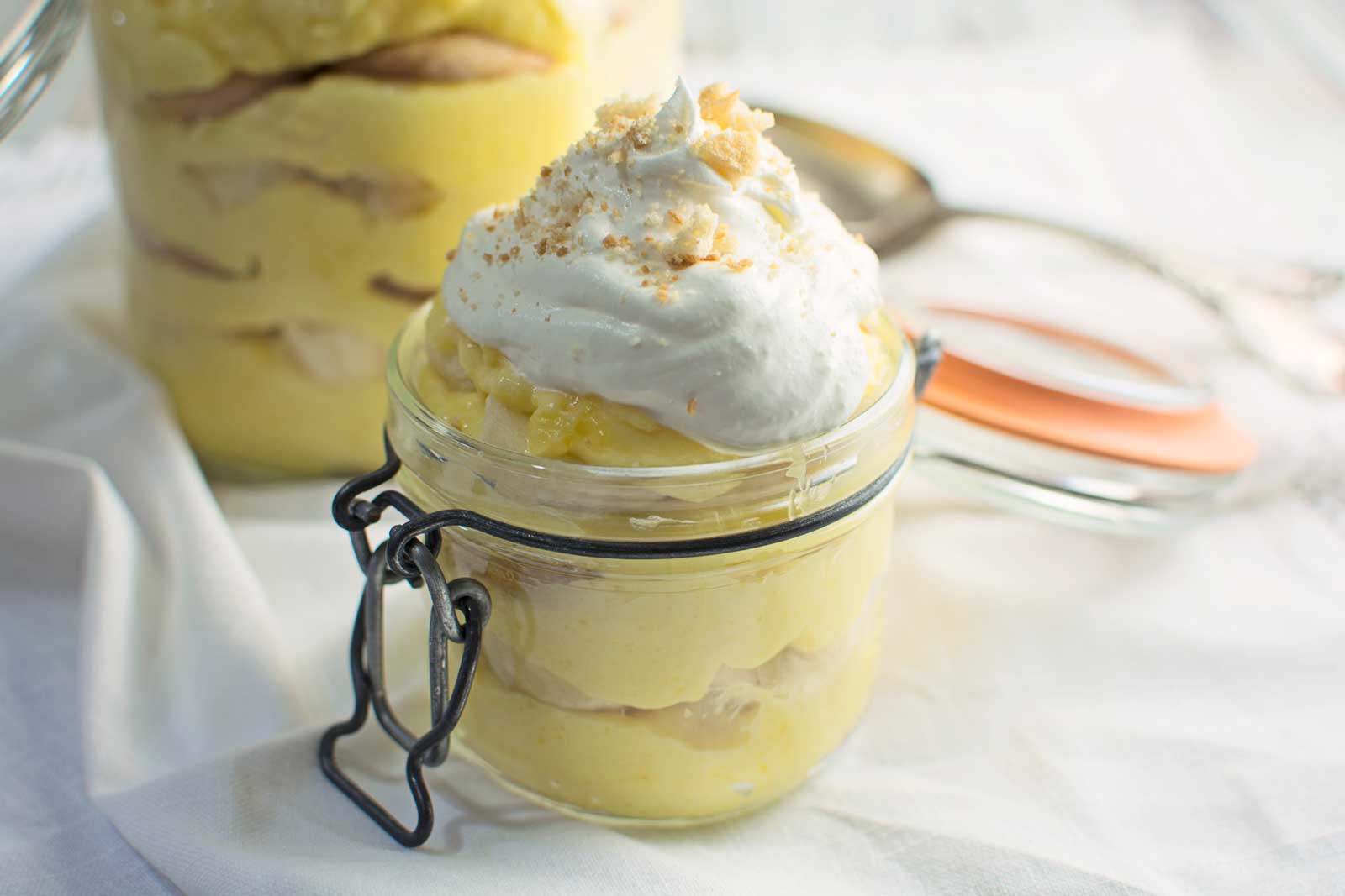 A small glass jar filled with layered Banana Pudding, topped with whipped cream and crumbled cookie pieces, sits on a white cloth with a spoon and jar lid in the background.