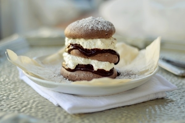 A Chocolate Pavlova dessert of three chocolate meringue layers filled with whipped cream and chocolate ganache, dusted with powdered sugar, sits on a white plate with parchment paper and a folded napkin underneath.