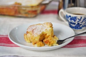 A slice of powdered sugar-topped Cream Cheese Coffee Cake sits on a white plate with a fork. In the background, theres a cup of coffee in a blue and white cup and a glass baking dish, all set on a white tablecloth with red stripes.