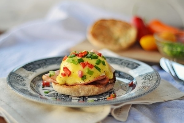 A plate with classic Eggs Benedict on an English muffin, topped with creamy hollandaise sauce, diced red and green peppers, and sliced ham, set on a patterned plate with vegetables blurred in the background.