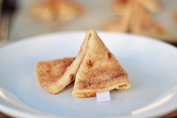 A close-up of a fortune cookie made from baked dough, sprinkled with cinnamon sugar, sitting on a white plate. A small white fortune slip is visible poking out from one side.
