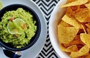 A bowl of fresh guacamole garnished with lime slices sits next to a bowl of yellow tortilla chips, all arranged on a black and white zigzag-patterned surface.