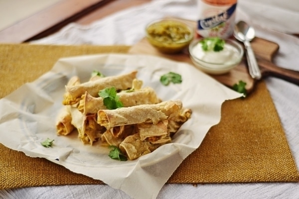 A plate of crispy rolled Chicken Taquitos garnished with cilantro, served on parchment paper. In the background are small bowls of green salsa and sour cream on a wooden board.