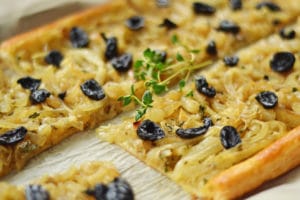 A Caramelized Onion Tart, this rectangular flatbread is topped with sweet caramelized onions, black olives, and fresh thyme, then sliced to reveal a golden, flaky crust.