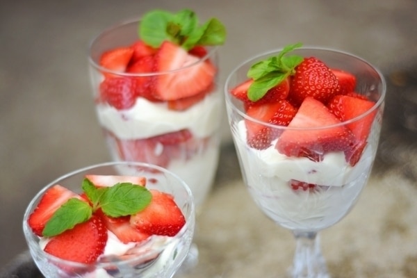 Three glass cups filled with layers of strawberries and cream parfait—whipped cream, sliced strawberries, and a mint sprig on top. The background is softly blurred for a dreamy effect.