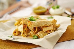A plate of rolled and baked chicken taquitos is arranged on parchment paper, garnished with fresh cilantro. In the blurred background, bowls of salsa and sour cream are visible.