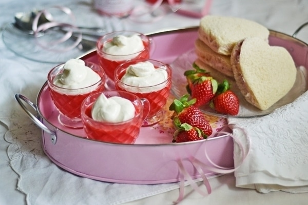 A pink tray holds four small cups of strawberries and cream jelly topped with whipped cream, fresh strawberries, and two heart-shaped sandwiches on a white tablecloth.