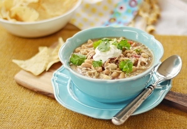 A comforting bowl of White Chicken Chili topped with sour cream and cilantro sits on a blue plate with a spoon. Tortilla chips are on the side, and a bowl of chips is blurred in the background.