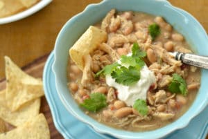 A blue bowl filled with White Chicken Chili, topped with sour cream and fresh cilantro. A tortilla chip is dipped into the chili, with more chips on the side. A spoon rests in the bowl, ready to enjoy this hearty dish.