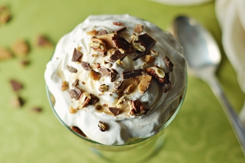 A glass dish filled with creamy whipped topping, sprinkled with chopped chocolate and nuts, sits on a green patterned surface with a spoon and scattered nut pieces nearby.