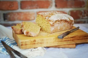 A loaf of rustic Irish brown bread sits on a wooden cutting board, with two slices cut and placed nearby. A knife rests beside the bread, and a blue-striped cloth is partially visible. A brick wall is in the background.