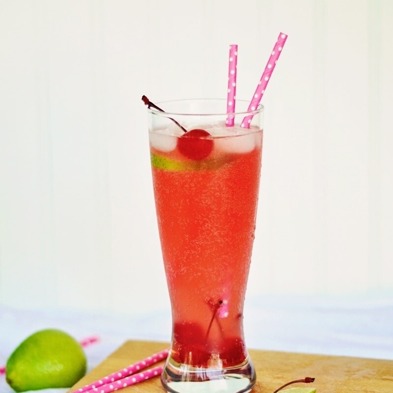 A tall glass of Cherry Limeade with ice, a cherry, and two pink polka-dot straws. Lime slices, a cherry, and a whole lime rest on a wooden board in front of the refreshing drink.