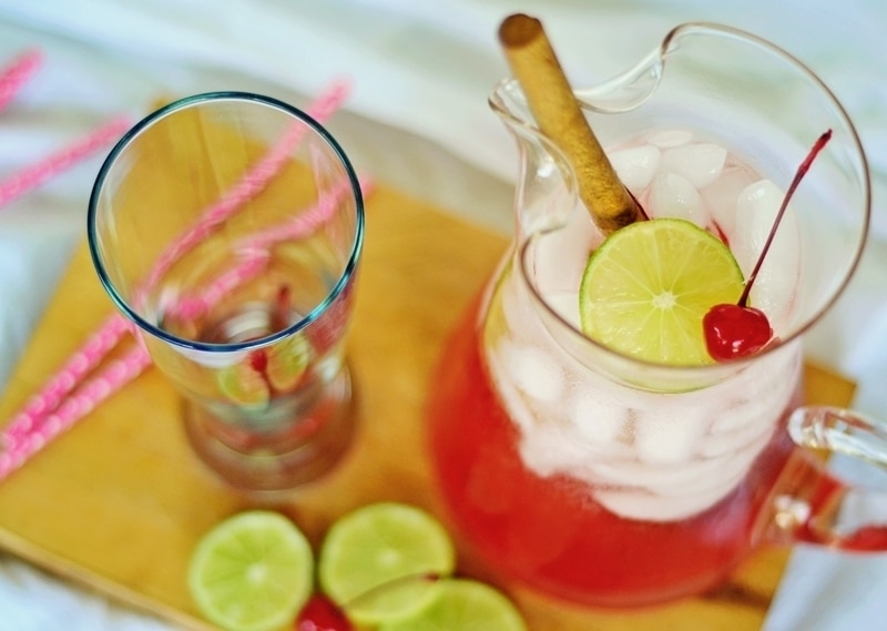 A pitcher of Cherry Limeade, garnished with a lime slice, cherry, and cinnamon stick, sits on a wooden board next to an empty glass, lime slices, and pink-striped straws.