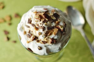 A glass dish filled with creamy whipped topping, sprinkled with chopped chocolate and nuts, sits on a green patterned surface with a spoon and scattered nut pieces nearby.