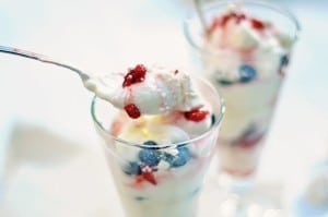 Two tall glasses filled with layers of creamy yogurt, blueberries, and raspberries create an Eton Mess Parfait. A spoon holds a scoop of yogurt and berries above one glass, highlighting the dessert’s fresh ingredients.