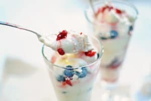 Close-up of a spoonful of creamy dessert with whipped topping, blueberries, and red berries above an Eton Mess Parfait glass layered with cream and berries. Another parfait-filled glass appears in the background.