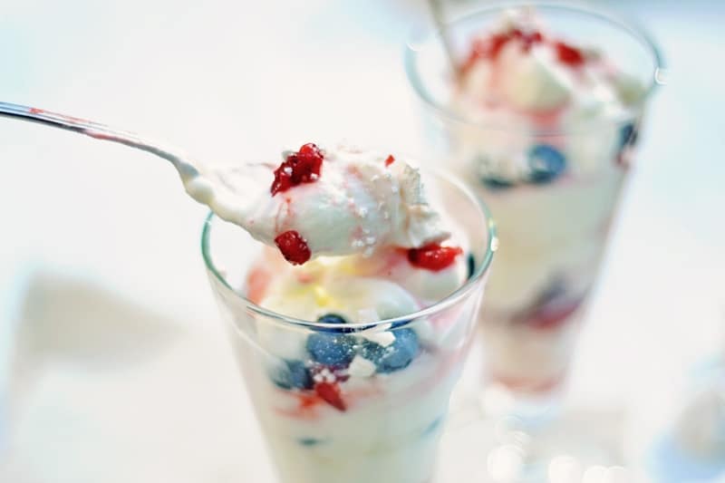 Close-up of a spoonful of creamy dessert with whipped topping, blueberries, and red berries above an Eton Mess Parfait glass layered with cream and berries. Another parfait-filled glass appears in the background.