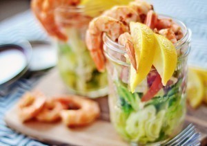 Two glass jars filled with layered salad ingredients, topped with shrimp cocktail and lemon wedges, are placed on a wooden board, with extra shrimp cocktail on the side.