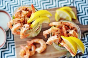 Two glass jars filled with Shrimp Cocktail, garnished with lemon wedges and crispy bits, sit on a wooden board, accompanied by extra shrimp and lemon slices on a patterned tablecloth background.