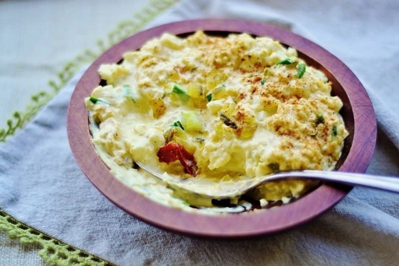 A wooden bowl filled with creamy potato and egg salad, garnished with sliced green onions and paprika, with a spoon resting inside. The bowl sits on a grey cloth napkin with a green crocheted edge.
