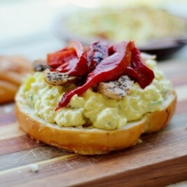 An open-faced sandwich with creamy egg salad, sliced grilled chicken, and strips of roasted red pepper rests on a wooden cutting board, with a bun top in the background.
