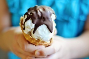 A person holds a waffle cone with coconut ice cream, topped with a hard chocolate shell. The person is wearing a bright blue shirt.