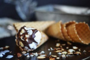 A close-up of a waffle cone filled with coconut ice cream, topped with chocolate sauce and chopped nuts, lying on a dark surface with scattered nuts and three empty cones in the background.