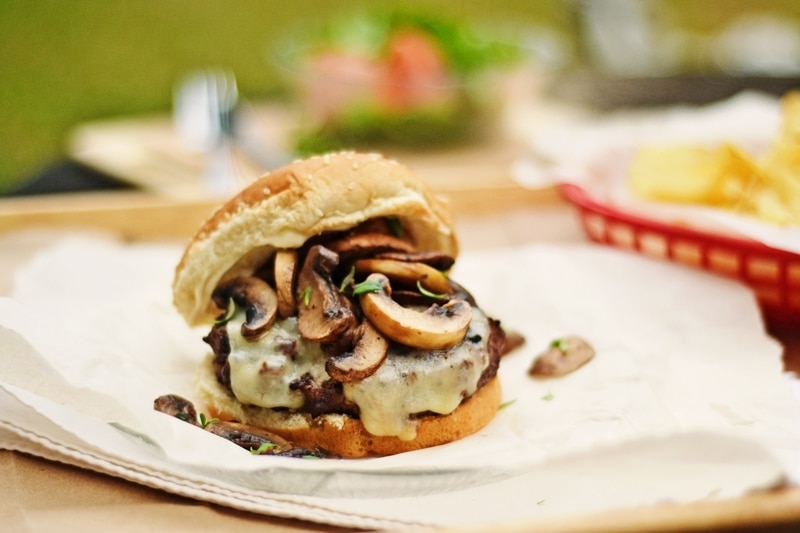 A close-up of a Mushroom Burger topped with sautéed mushrooms on a paper-lined tray, with a blurred background showing a basket of chips and a bowl of salad—perfect inspiration for your next mushroom burger recipe.
