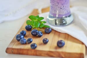 Fresh blueberries and a sprig of mint are scattered on a wooden board next to a glass of blueberry smoothie, creating an inviting scene with a white napkin in the background.