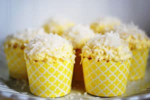 A group of Coconut Cupcakes in yellow patterned wrappers, topped with yellow frosting and shredded coconut, arranged on a glass plate.
